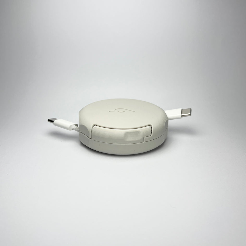 the bottom of the off-white Pebbl 1 mini, with the pebbl Innovations Ltd logo at the bottom of an automatic cable organiser with official Apple 1m charging cable, designed to retract cables instantly at the click of a button.